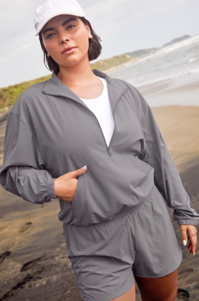 model is wearing a gray stretch woven quarter zip jacket with a kangaroo pocket and gray stretch woven shorts. 