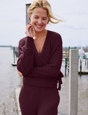 A woman smiling outdoors near a waterfront dock, wearing a matching burgundy knit wrap top and pants set.