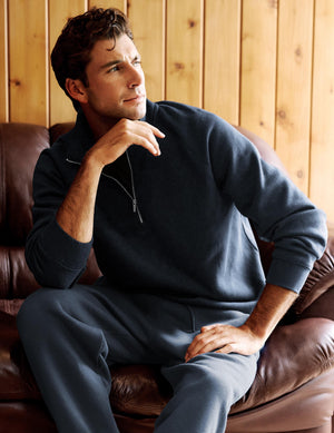 A man wearing a dark navy blue zip-up sweatshirt and matching sweatpants sits on a brown leather couch in front of a wood-paneled wall.