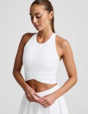 Woman wearing a white ribbed high neck cropped tank and a white skirt against a plain background. 
