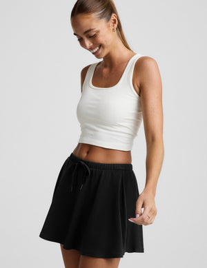 Woman wearing a white tank top and black skirt on a plain background. 