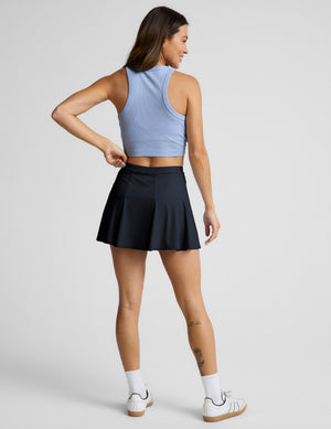 Woman wearing a light blue ribbed tank top and navy blue mini athletic skirt against a plain background. 