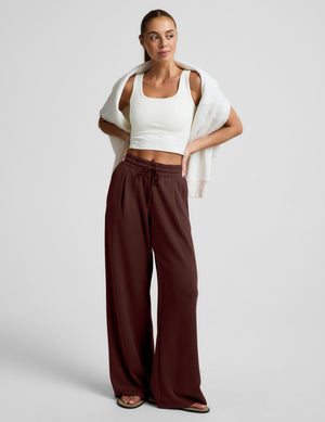 Woman wearing a white cropped tank, brown wide leg pants, and a white sweatshirt draped over her shoulders on a plain background. 