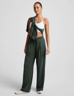 Woman wearing a white bra and green wide leg pants against a plain background. 