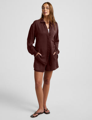 Woman wearing a brown button up shirt and shorts against a plain background. 