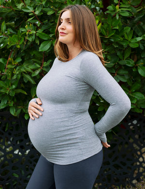 A pregnant woman wearing a light gray long-sleeve maternity top and dark blue leggings, posing outdoors in front of a green leafy hedge.