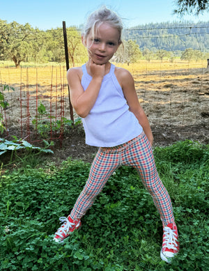 Toddler wearing a white tank top and gingham printed leggings standing on grass. 