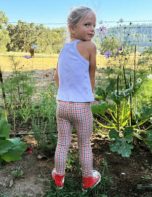 Toddler wearing a white tank top and gingham printed leggings standing on grass. 