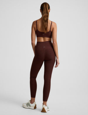 Woman wearing a brown sports bra and brown high-waisted midi length leggings against a plain background. 