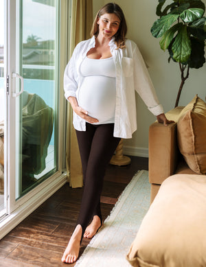 A pregnant woman stands barefoot in a living room doorway, wearing a white button-up shirt over a white maternity tank top, dark brown leggings, and tan boots. She is holding her baby bump and leaning on a tan sofa next to a large green plant.