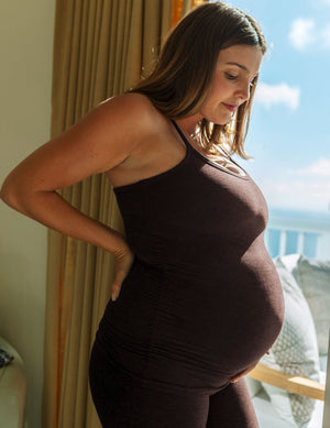 Pregnant woman wearing a brown racerback tank top and matching maternity leggings standing in front of a window. 