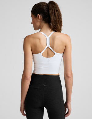 A smiling woman wearing a white crop top tank with thin shoulder straps, paired with black bottoms, photographed against a light gray studio background.