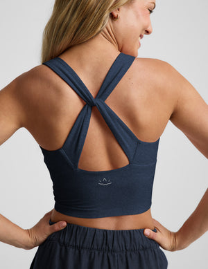 A woman shown from the back wearing a navy blue cropped tank with a crossover crisscross strap design and a small white logo on the lower center back, paired with matching dark bottoms against a light gray studio background.