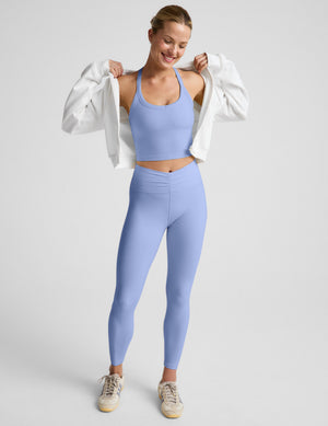 Woman wearing a light blue cropped tank top and leggings with a white zip-up sweatshirt against a plain background. 