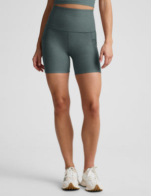 Woman wearing green biker shorts with pockets against a plain background. 