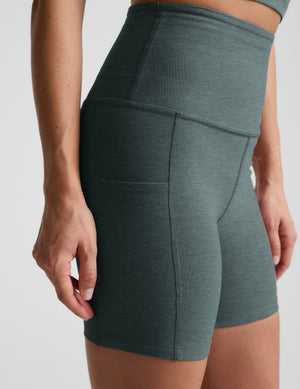 Woman wearing green biker shorts with pockets against a plain background. 