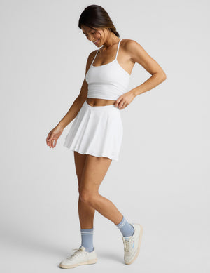 Woman wearing a white cropped tank top and athletic skirt with built-in shorts with pockets against a plain background. 