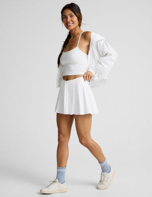 Woman wearing a white cropped tank top and athletic skirt with built-in shorts with pockets against a plain background. 