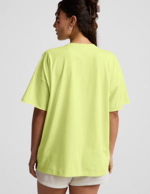 Woman wearing a bright level v-neck oversized t-shirt and white shorts against a plain background. 