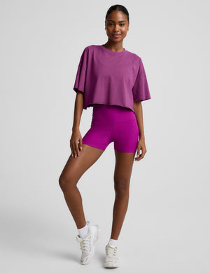 Woman wearing a purple cropped t-shirt and purple biker shorts against a plain background. 