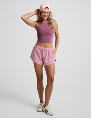 Woman wearing a purple ribbed cropped tank and pink fleece shorts against a plain background. 