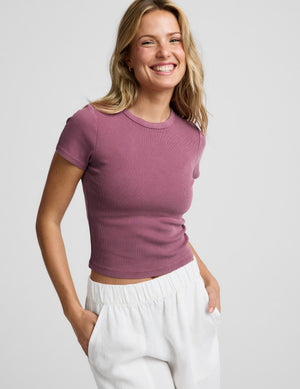 Woman wearing a purple ribbed baby tee and white linen pants against a plain background. 