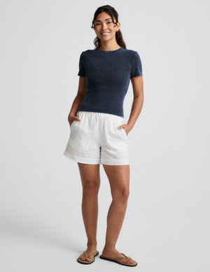 Woman wearing a navy blue ribbed t-shirt and white linen shorts against a plain background. 