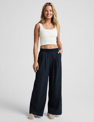 Woman wearing a white ribbed tank top and blue pants against a plain background. 