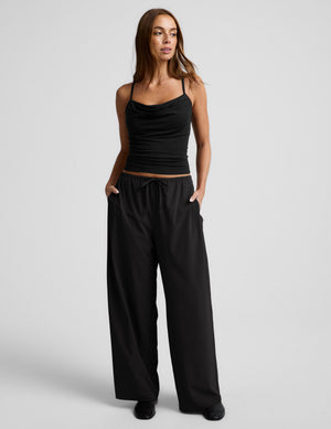 Woman wearing a black tank top and wide-leg pants on a light gray background. 