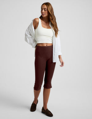 Woman wearing a white tank top and brown pedal pushers on a plain background. 