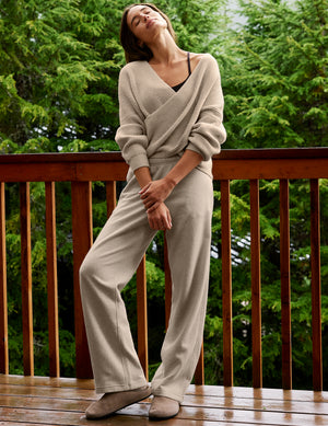 A woman wearing a beige knit wrap sweater and matching wide-leg pants stands on a wooden deck with green trees in the background.