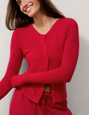 A woman wearing a bright red long-sleeve button-front cardigan and matching red pants, photographed against a plain light gray wall.