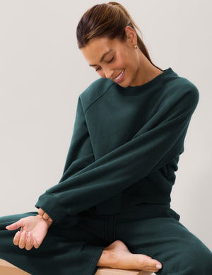 A woman wearing a dark forest green crewneck sweatshirt and matching sweatpants, sitting cross-legged against a plain light gray background.