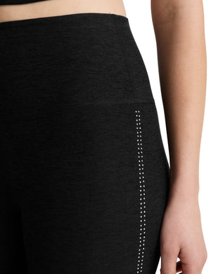 A woman modeling a black athletic set consisting of a cropped tank top and high-waisted leggings, standing against a light gray studio background.