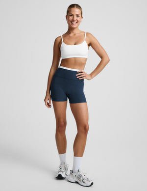 Woman wearing a white sports bra and navy blue biker shorts against a plain background. 