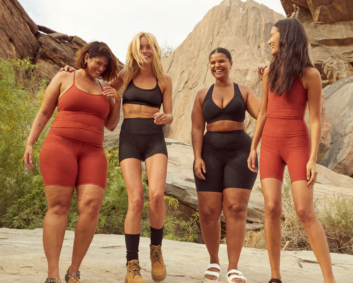 Four women standing and laughing in shorts and sports bras hiking
