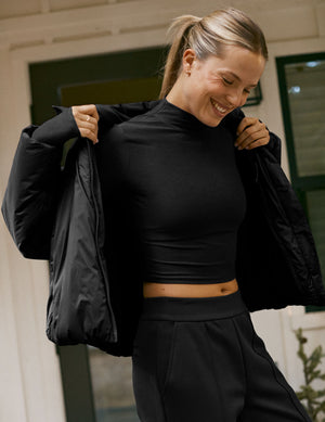 Woman wearing a black puffer jacket over a black top and pants, standing indoors.