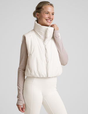 model wearing a white puffer vest over a long-sleeve shirt on a light gray background. 