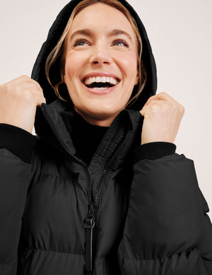 woman wearing a long black hooded puffer jacket on a light background. 