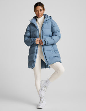 Woman wearing a blue mid length puffer coat on a light gray background. 