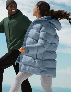 woman wearing a blue puffer coat standing against a mountainous landscape with a clear sky next to a man in a green jacket and black pants. 