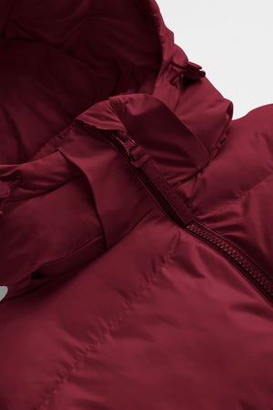 woman wearing a red mid thigh length puffer jacket with a hood against a snowy background. 