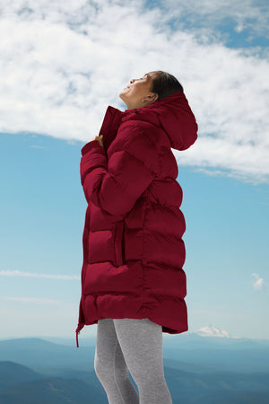 woman wearing a red mid thigh length puffer jacket with a hood against a snowy background. 