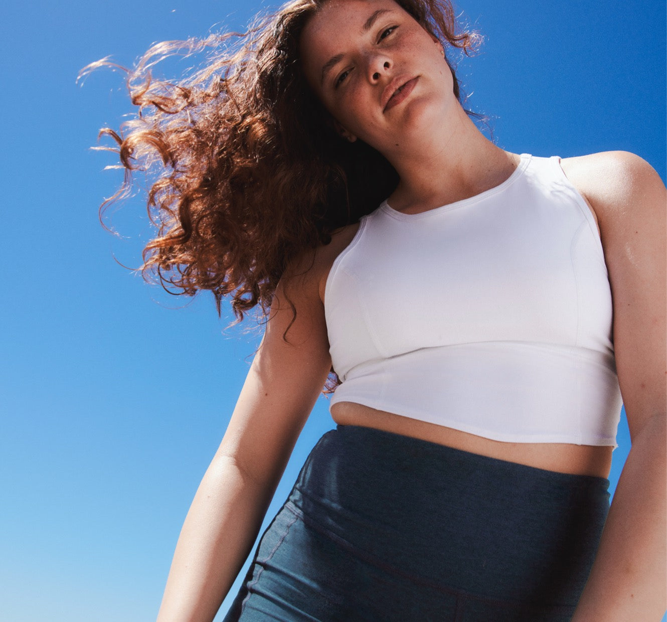 model is wearing a white high-neck cropped tank top and blue high-waisted midi leggings. 