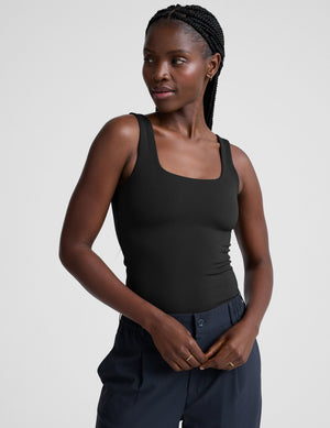 model is wearing a black double-layered square neckline bodysuit and blue trousers against a light gray background. 