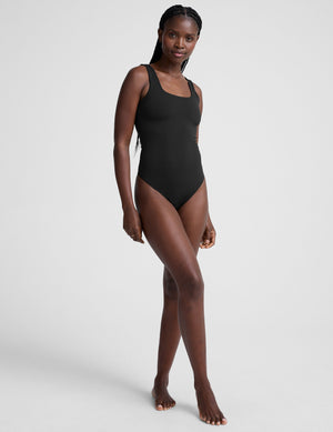 model is wearing a black double-layered square neckline bodysuit and blue trousers against a light gray background. 