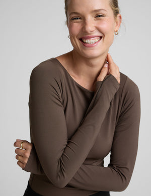 woman wearing a brown long-sleeve boat neck bodysuit and black pants against a plain background. 