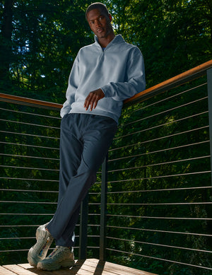 Man in a forest leans on a balcony wearing navy Dynamic Men's Straight Leg Pant and a light blue sweatshirt.