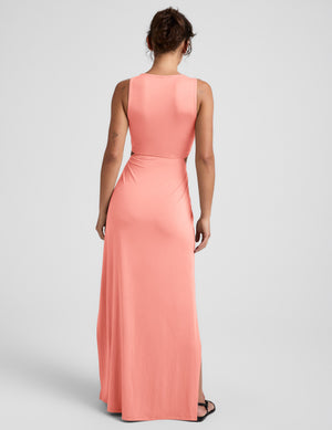 Woman wearing a coral pink maxi dress with cut-out design on a light gray background. 