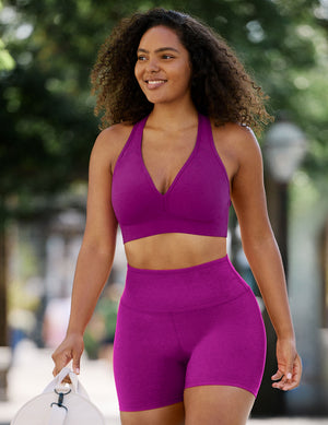 A woman wearing a matching magenta-purple activewear set consisting of a halter-neck sports bra and high-waisted biker shorts, walking outdoors on a blurred street background.